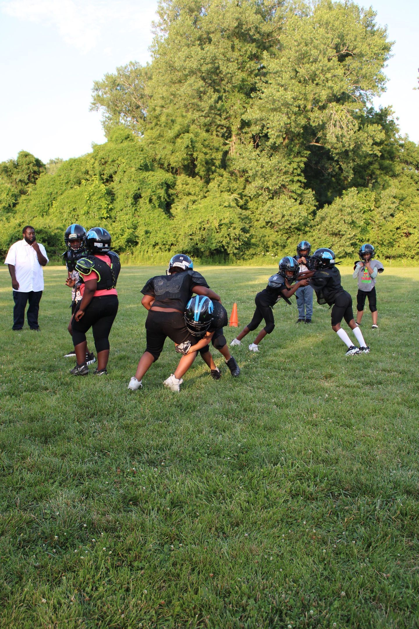 Youth football players in black uniforms practicing on a grassy field with a coach watching.