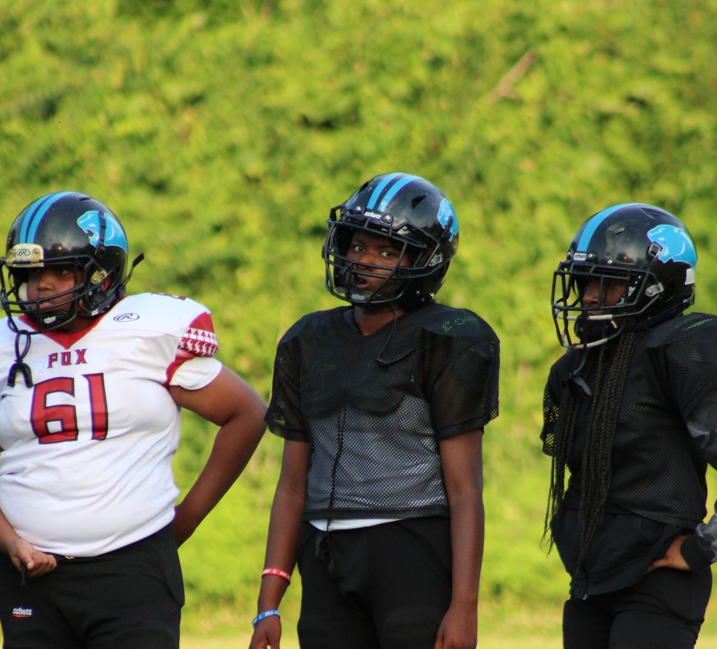 Three youth football players in helmets and pads stand outside on a green field.