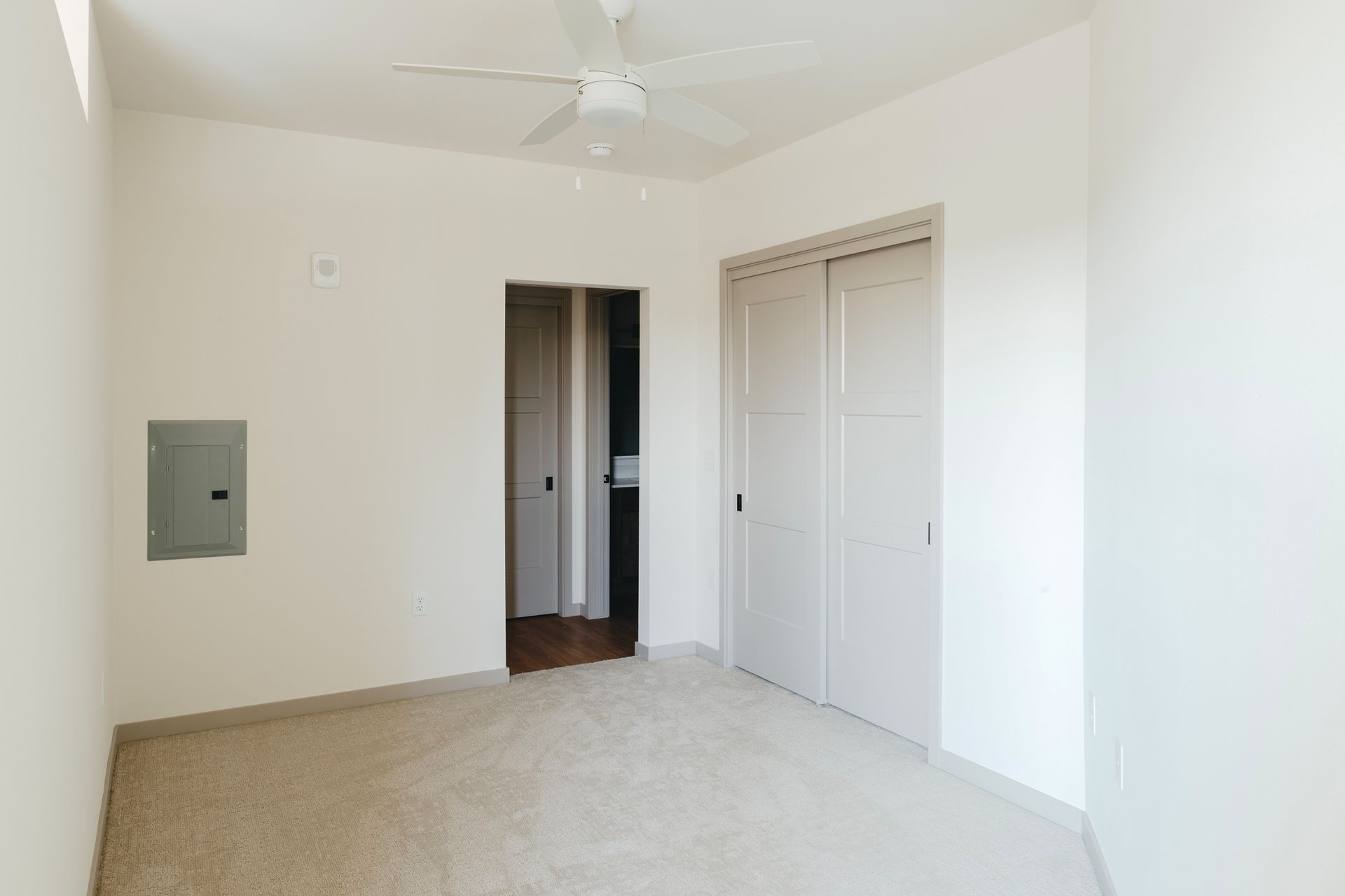 Empty white-walled bedroom with closet and open doorway to another room; beige carpet and ceiling fan at Revelry in Madison WI.