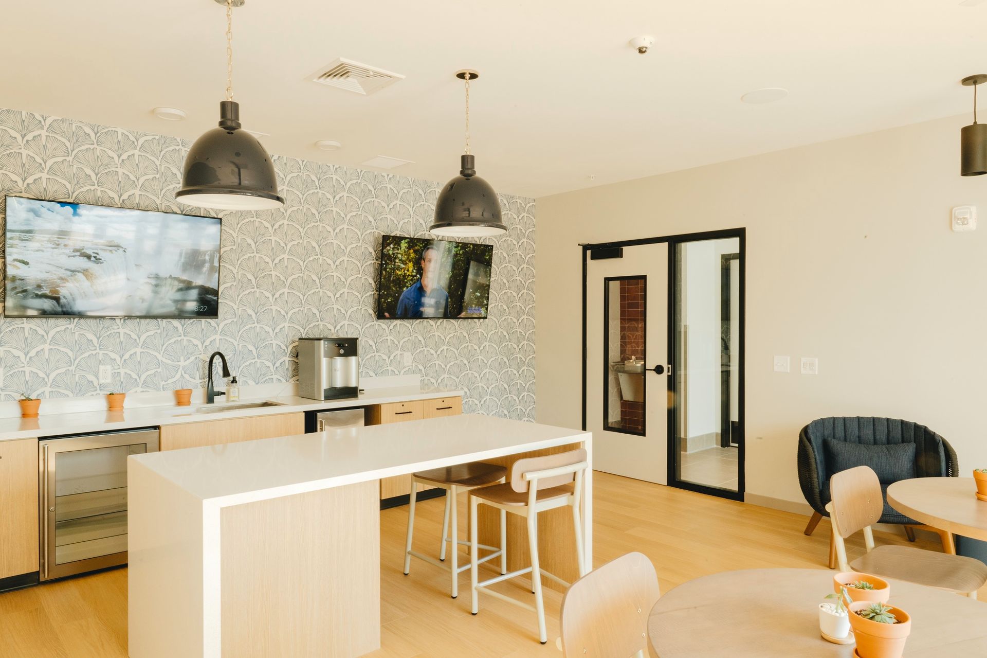 Kitchen area with two TVs, a bar, and seating. The space has light wood floors, and a patterned wallpaper at Revelry in Madison, WI. 