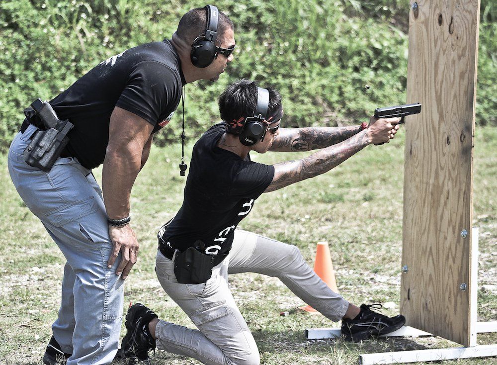 Man coaching person kneeling, aiming handgun at target in outdoor setting. Both wear ear protection.