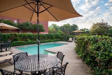 Poolside scene with umbrella, table, chairs, lounge chairs, pool, and green hedges under a cloudy sky.