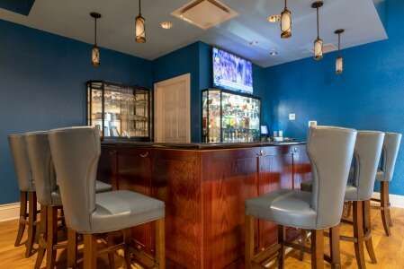 A bar area with blue walls, wooden bar, bar stools, and hanging lights; a TV is mounted on the wall.