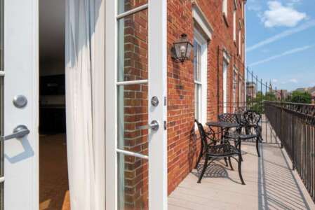 Open white French doors lead to a balcony with a black wrought-iron table and chairs on a brick building.
