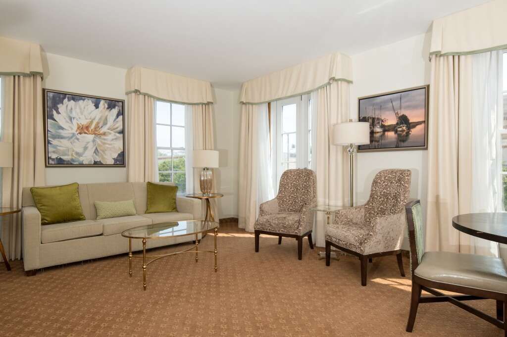 Living room with sofa, chairs, windows, and artwork. Beige walls, carpet, and light-colored curtains.