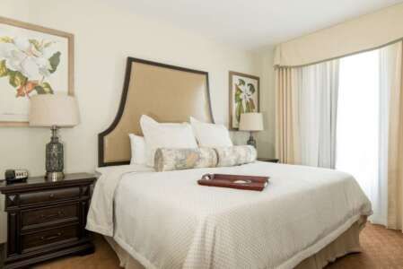 Bedroom with a large bed, two lamps, framed art, and a window with curtains; beige and brown tones.