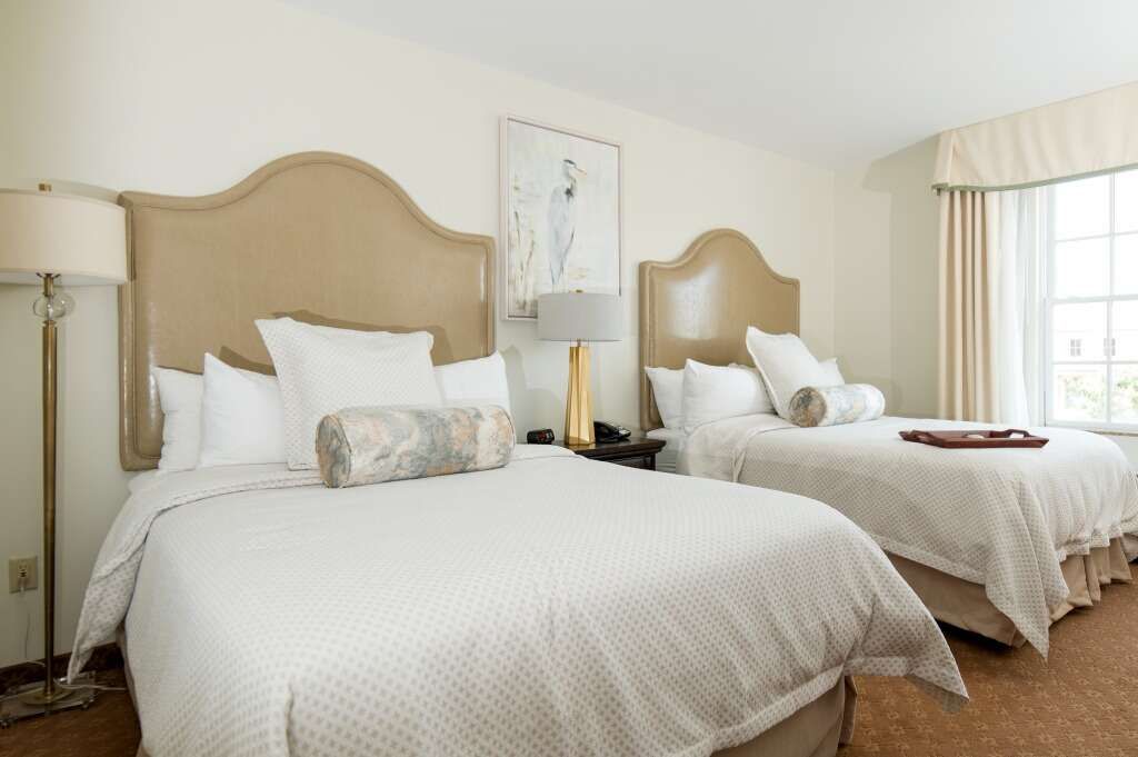Hotel room with two queen beds, beige headboards, white bedding, and a window with a curtain.