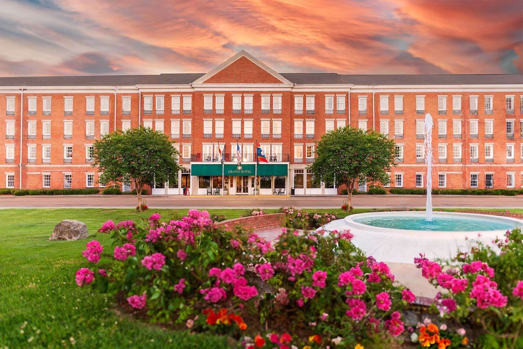 Brick hotel with fountain and pink flowers against sunset sky.