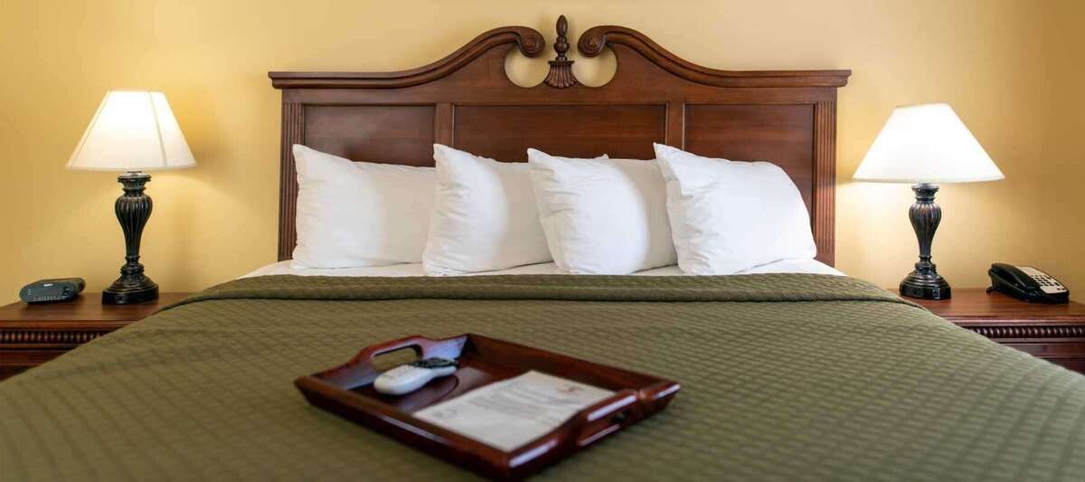 A hotel bed with a wooden headboard, white pillows, and green blanket. A tray with remotes sits on top.