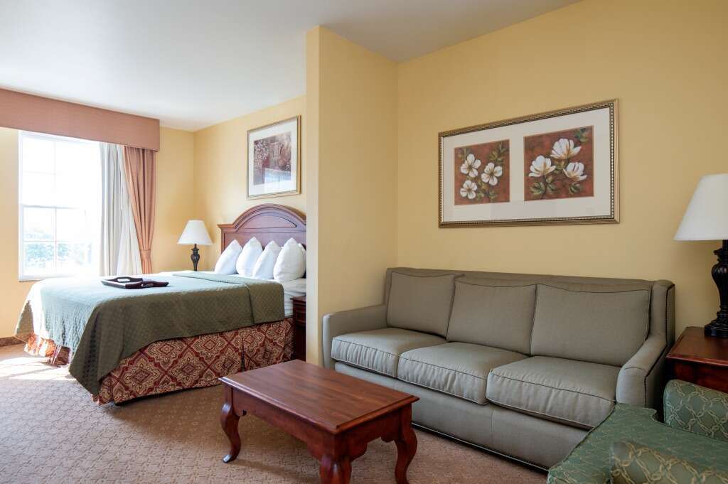 Hotel room with bed, sofa, and small table. Yellow walls, green bedspread, beige carpet.