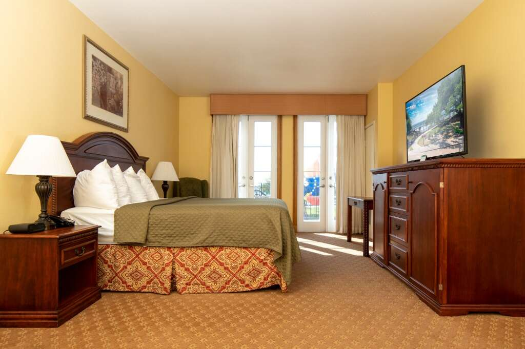 Hotel room with a bed, TV, dresser, nightstand, and balcony doors. Yellow walls and brown furniture.