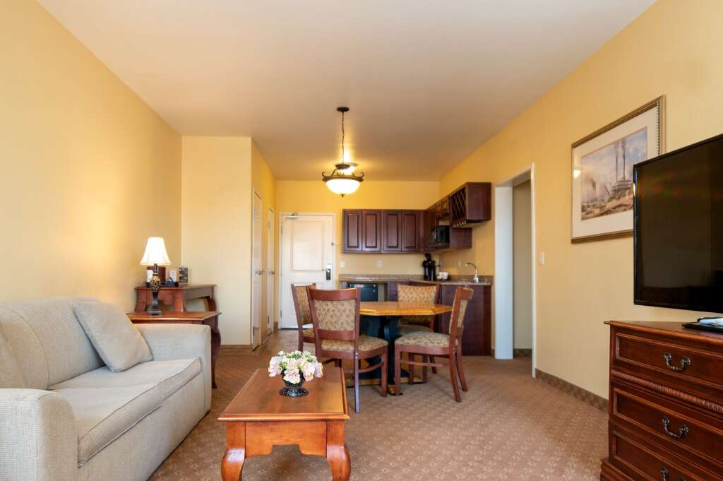 Cozy hotel suite with a sofa, dining area, and kitchenette; warm tones, wood furniture.