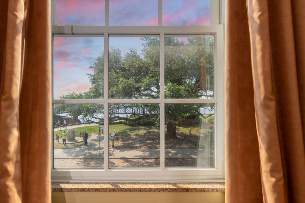 Window view of a tree and landscape, with a pink sky, framed by brown curtains.