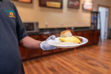 Person in black shirt and gloves holds a plate of food: grits, eggs, biscuit, and sausage.