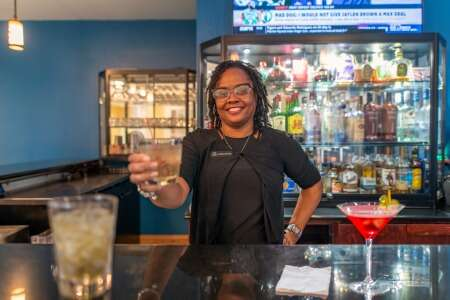 Bartender smiles, holding a drink over a bar; bottles and drinks in the background.