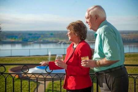 Elderly couple on a balcony overlooking a river, holding champagne glasses.