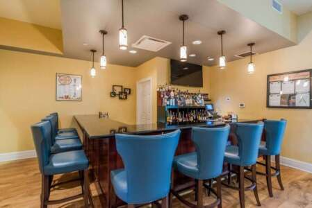 Bar with blue chairs, dark countertop, bottles, TV, and pendant lights.
