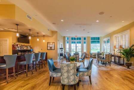 Lounge with bar, tables, and chairs. Light wood floors and blue and gold decor. Windows in the background.