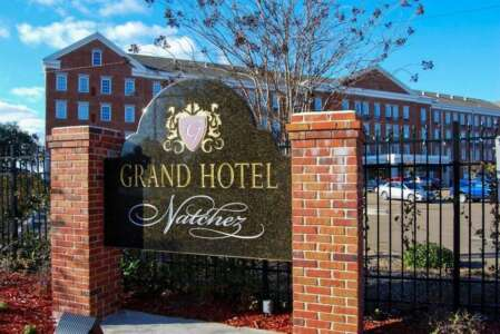 Sign for Grand Hotel Natchez with red brick pillars, a wrought iron fence, and the hotel in the background.