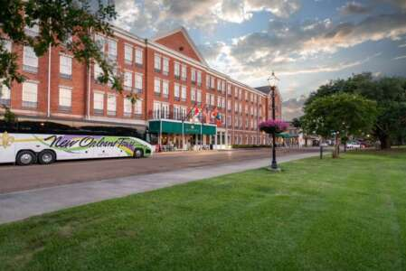 Red brick hotel with green awning entrance. A tour bus sits on the street. Green grass and trees in foreground.