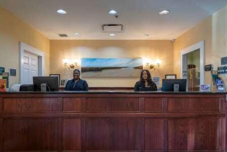 Two women at a hotel reception desk, smiling. Dark wood desk, neutral wall, artwork, and computers.
