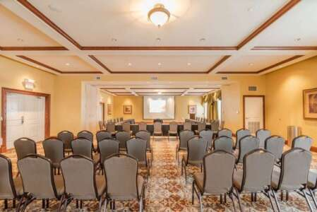 Conference room with rows of chairs facing a projector screen, beige walls, and ornate ceiling.