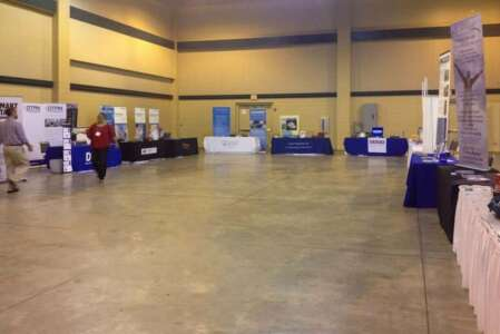 Exhibition hall with booths, people, and products displayed on tables. Beige walls and concrete floor.