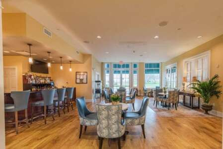 Bar and seating area in a light-filled room, with a bar, tables, chairs, and large windows.