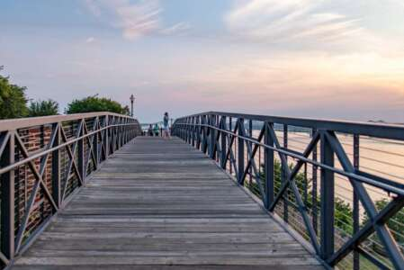 Wooden bridge over water, metal railings, people walking, sunset sky.