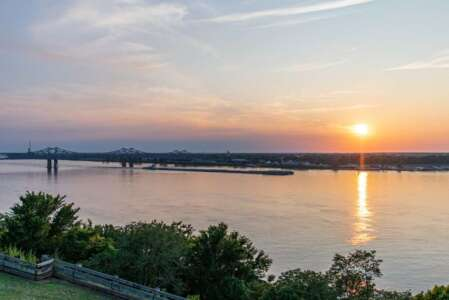 Sunset over wide river with bridge in silhouette; orange and pink hues, trees in foreground.