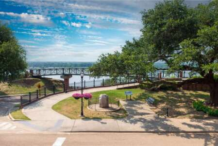 Park scene with a curved walkway, trees, and a view of a body of water under a cloudy sky.
