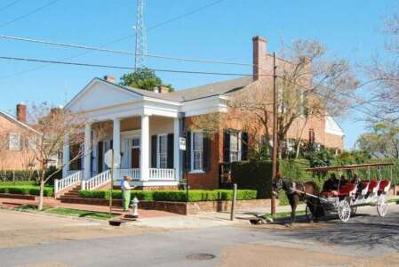 A brick building with white columns and a horse-drawn carriage on a sunny day.