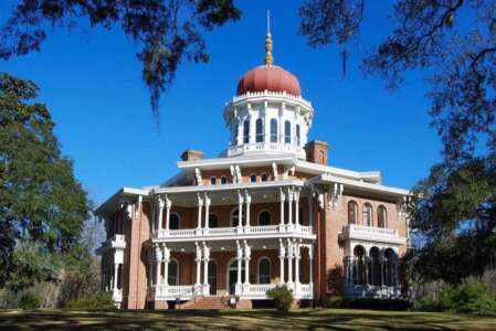 Large brick mansion with a red dome, multiple levels of balconies, and trees under a bright blue sky.
