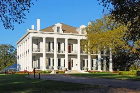 White, two-story plantation home with large columns, set on a green lawn under a clear blue sky.