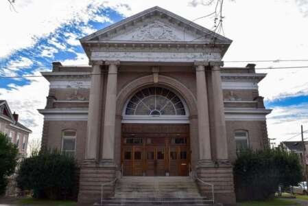 Classic building with columns, arched entryway, and steps leading to double doors. Light brown exterior, cloudy sky.