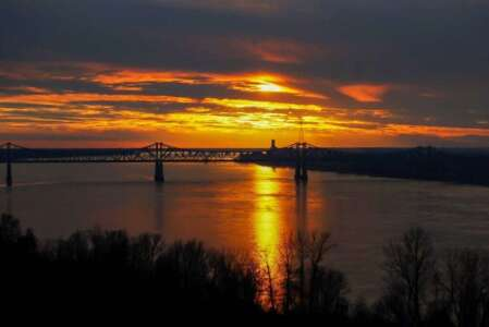 Sunset over a river with a bridge, reflecting orange and yellow hues. Silhouetted trees in foreground.