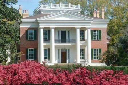 Red brick mansion with white columns, two stories, green shutters, and red azaleas in front.