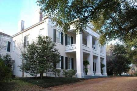 White two-story house with pillars, dark green shutters, and trees, on a grassy lawn.