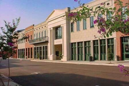 Row of beige and brick buildings with shops; street lined with trees and flowers.