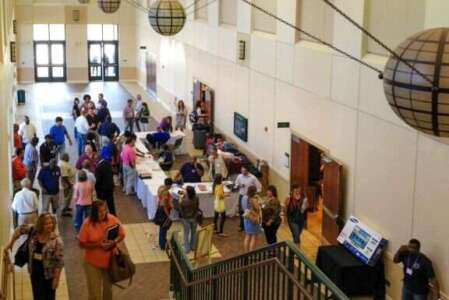 People at an event, standing and walking in a brightly lit hall. Several tables present information.