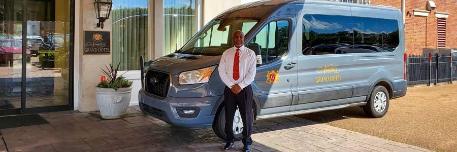 A man in a white shirt and red tie stands near a gray shuttle van in front of a hotel.