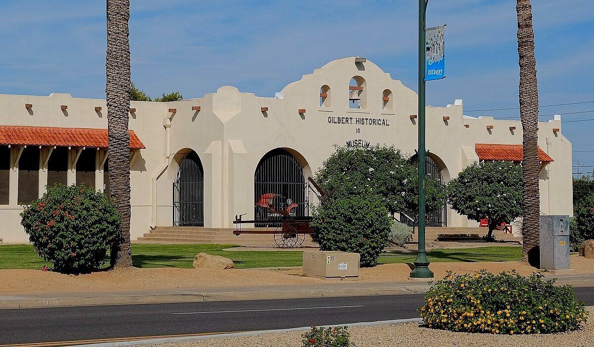Spanish-style building with stucco walls, arched windows, palm trees, and shrubs on a sunny street