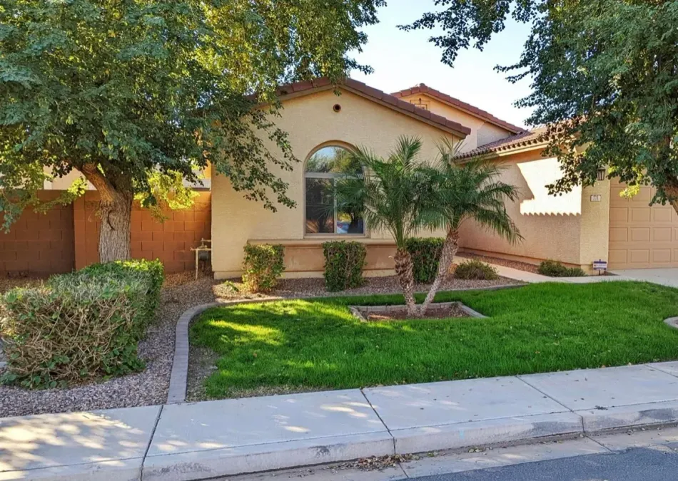 Single-story tan house with arched window, palm trees, green lawn, and a sidewalk in front