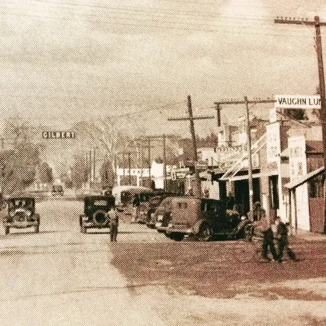 Old sepia street scene with early cars, storefronts, utility poles, and pedestrians on a dusty road.
