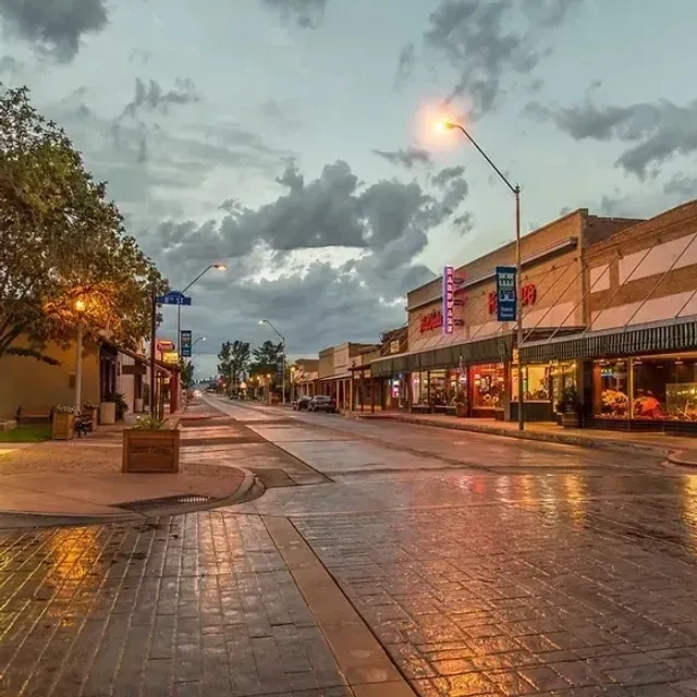Empty wet downtown street at dusk, with storefronts, glowing streetlights, and cloudy sky.