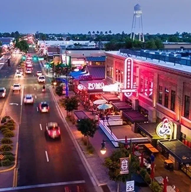 Evening downtown street with cars, lit shops, and a water tower in the distance