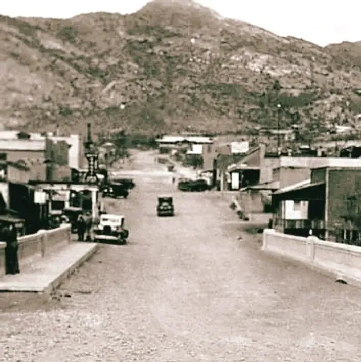 Old black-and-white street scene with cars on a bridge, low buildings, and mountains in the background