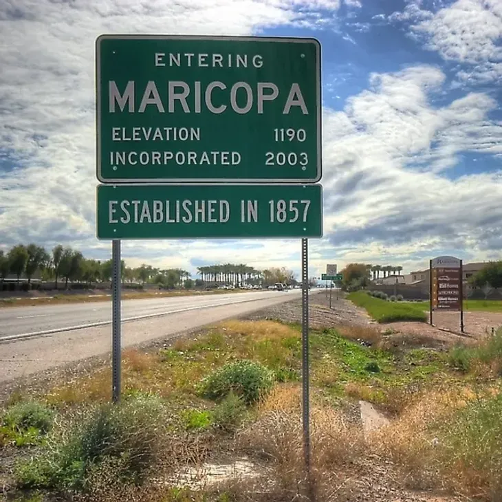 Roadside sign entering Maricopa, green with white text, beside a highway under a cloudy sky.