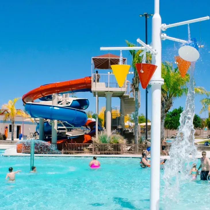 Outdoor water park with blue and orange slides, swimmers, and a splash fountain under a bright blue sky