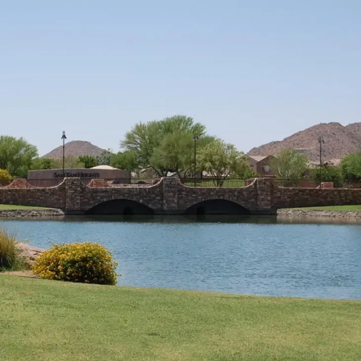 Stone bridge over a calm blue lake with green grass, shrubs, and desert hills in the background